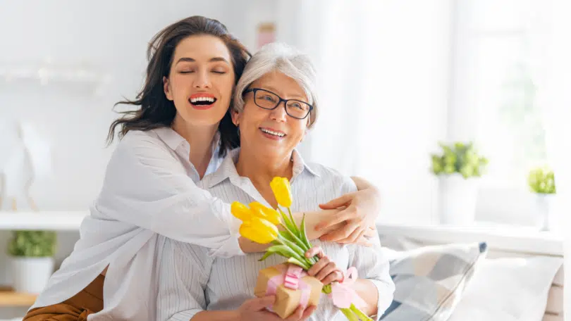 Duas mulheres se abraçando e sorrindo com flores e um presente nas mãos